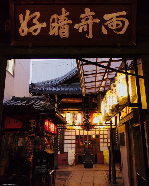 Un très vieux temple de Kyoto coincé entre des immeubles, on voit son enseigne ainsi que des lanternes qui éclairent son entrée et jusqu'au hall du temple